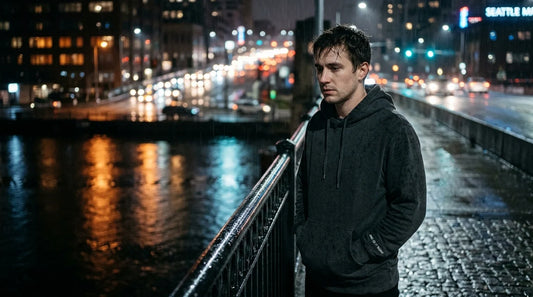 Man in dark embroidered hoodie on rainy bridge at night.